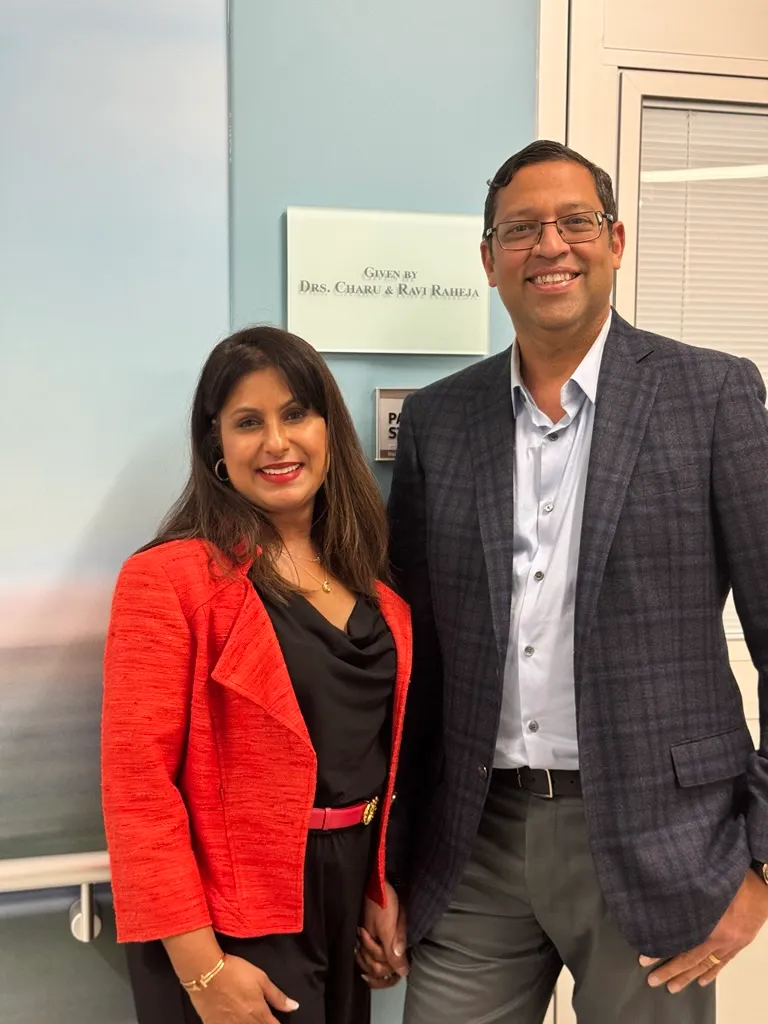 Two smiling people standing in front of a wall plaque that reads 'Given by Drs. Charu & Ravi Raheja'
