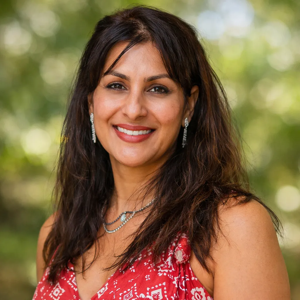 A woman with long dark hair smiling at the camera, wearing a red patterned top and drop earrings, against a blurred green outdoor background.