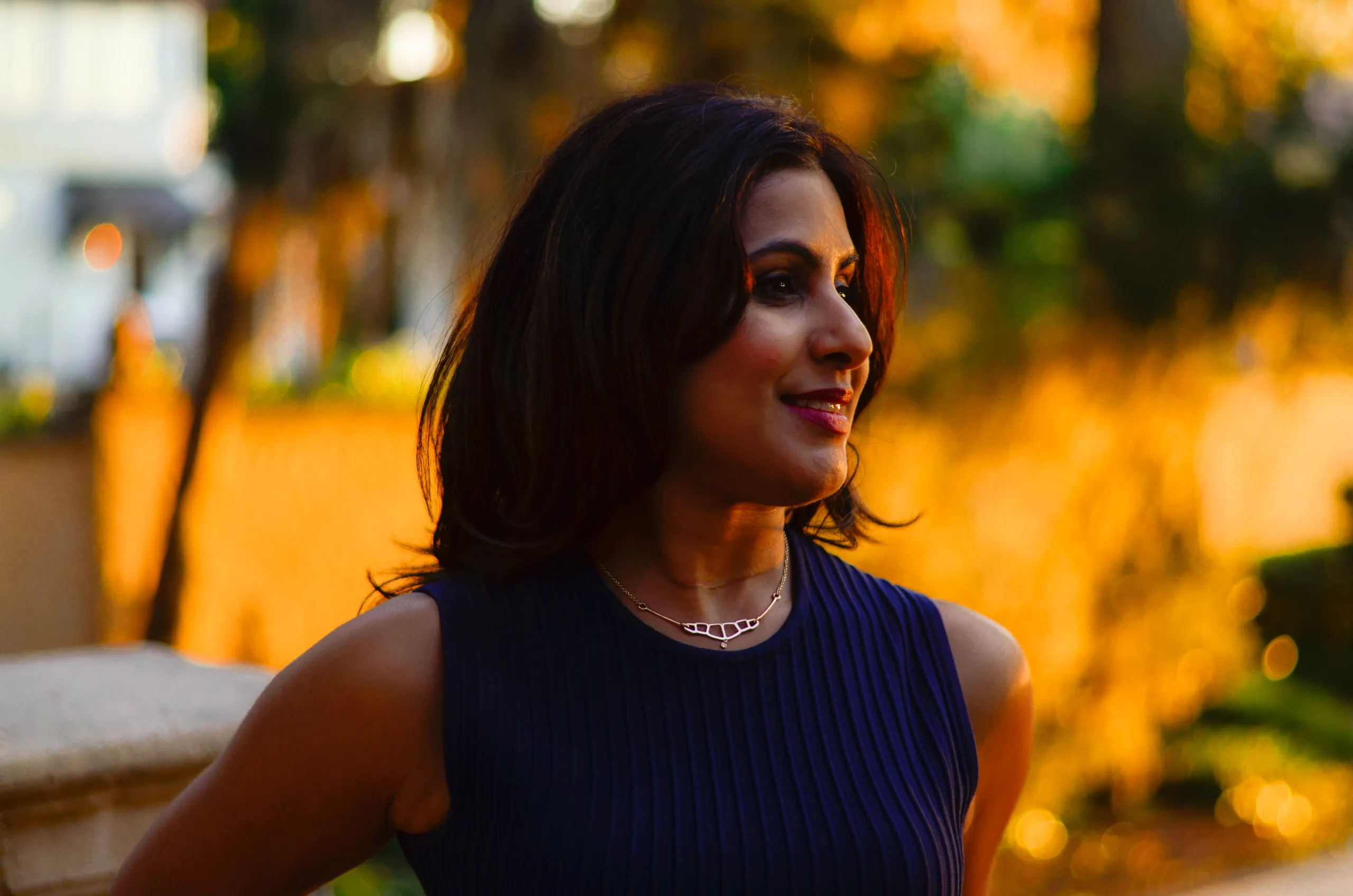 A woman in a navy sleeveless top looks off to one side in warm golden-hour light, with a blurred outdoor background behind her.