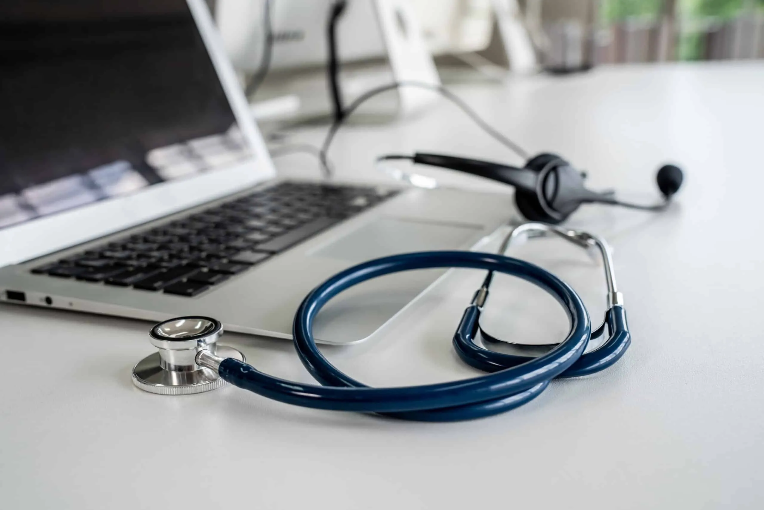 A blue stethoscope and a phone headset sit on a white desk in front of an open laptop